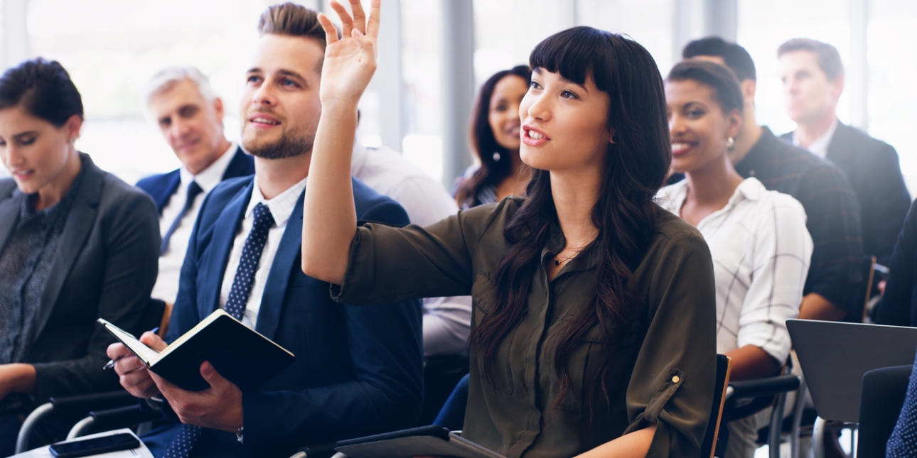 A group of business people of different ages, genders and ethnicities sat in a lecture. A woman closest to the camera raises her hand.