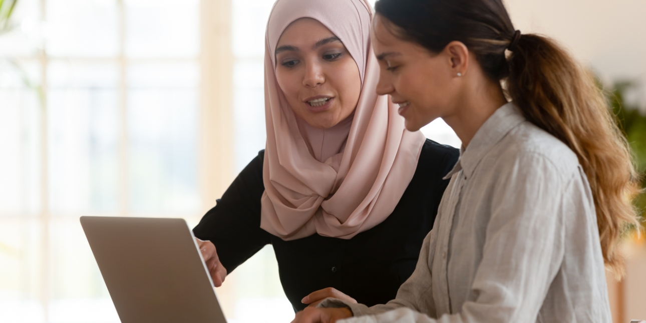 Two women sat around a laptop in a room lit by a large window. The woman on the left, who wears a pink hijab and black long sleeve top, points at the screen. The woman on the right, who has long brown hair tied in a ponytail, wears a long-sleeve grey