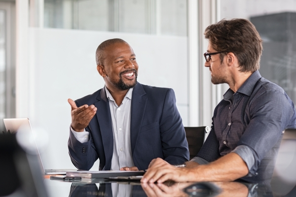 Two colleagues chatting over a desk. On the left is a black male wearing a navy suit jacket and grey scarf who is smiling. On the right, the side profile of a white male colleague wearing a navy shirt and glasses who is listening intently