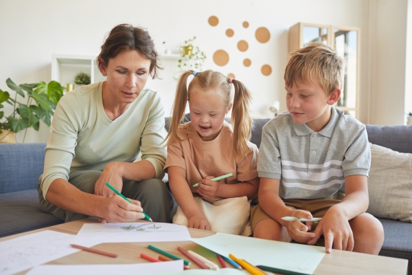 A mother, daughter and son are drawing together on a coffee table while sat on a sofa. The mother is white with dark hair and green top. The daughter is white, has blonde hair and has Down's syndrome. She wears a peach dress and is smiling. The son i