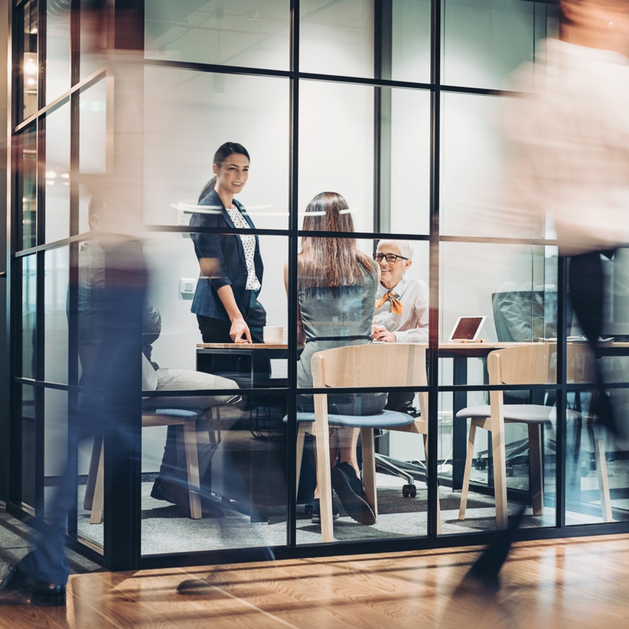 Three women inside a glass fronted meeting room. One is seen from the back and has long brown hair. The other two are white women, one sat and one stood up. The blurred outline of people walking past are in the foreground