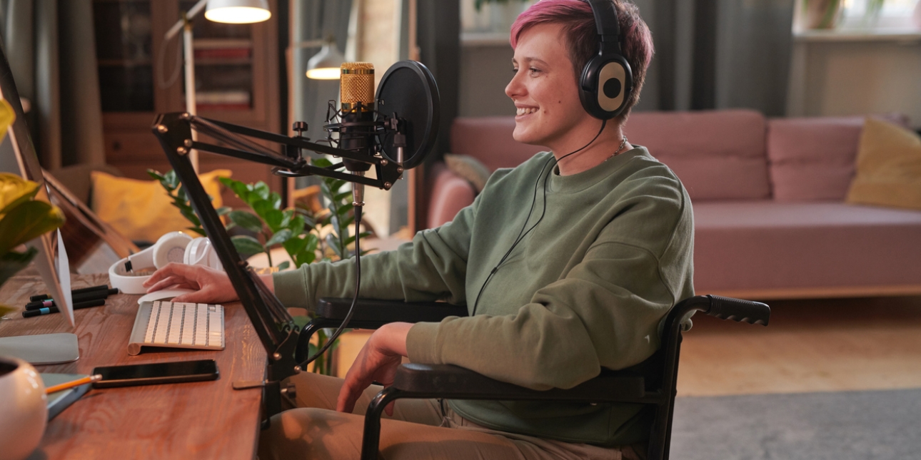 A white woman with short pink hair and wearing large headphones is sat in a wheelchair in front of a desk with podcasting equipment including a microphone