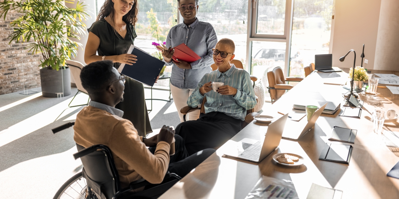 In a bright office, four smiling colleagues in smart-casual wear chat over coffee. A Black man who uses a wheelchair is speaking directly to a Black woman sat next to him, clutching a cup. Two other women - one white, one Black - are stood next to th