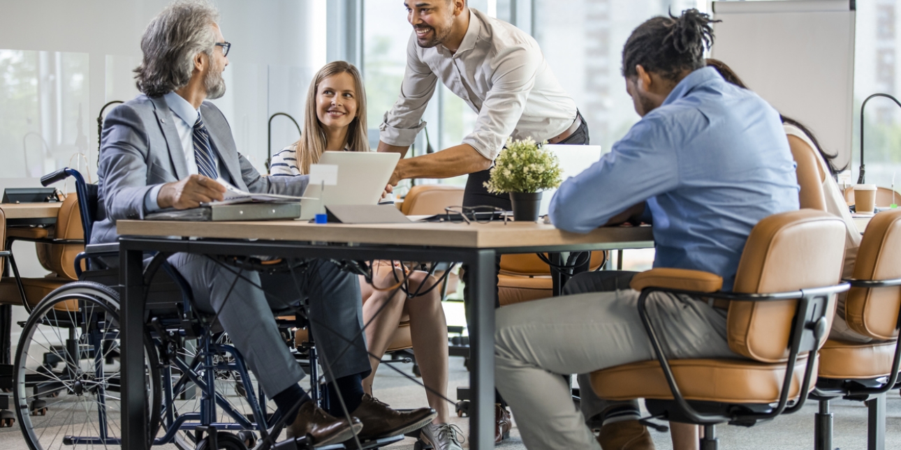 Five business colleagues sat around a table. On the left is a man sat in a wheelchair, next to him a woman smiling and stood over the desk is man also smiling. Another man and woman have their backs to camera.