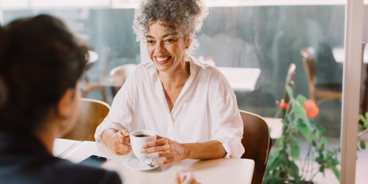 Happy mature businesswoman smiling while having a discussion with her business partner. She is holding a cup of coffee.