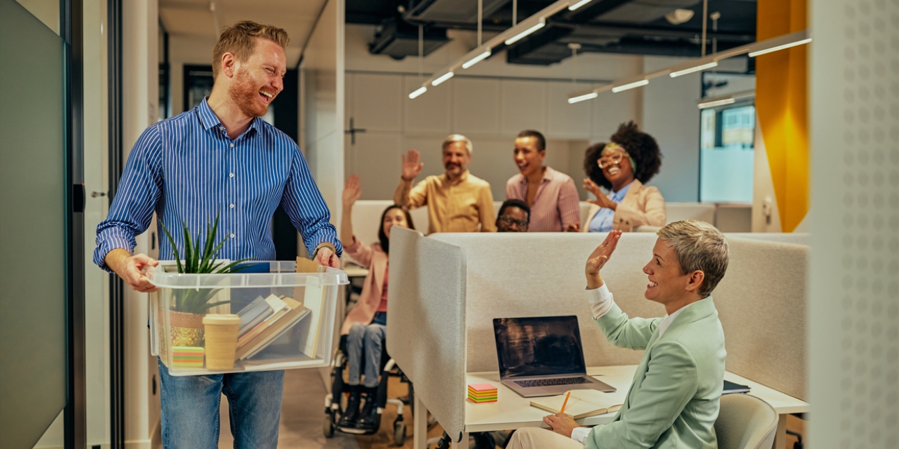 A man, who is white with ginger hair, carries a box of belongings from the office, smiling at six colleagues who are waving goodbye