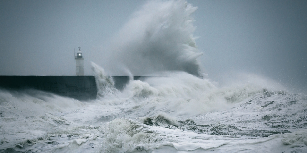 Large waves break against a stone jetty and rise into the sky. In the background is a lighthouse. The sky is a dark grey.