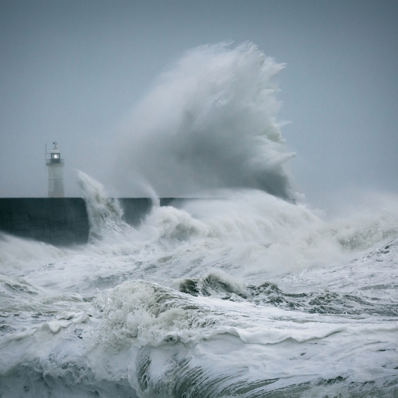 Large waves break against a stone jetty and rise into the sky. In the background is a lighthouse. The sky is a dark grey.