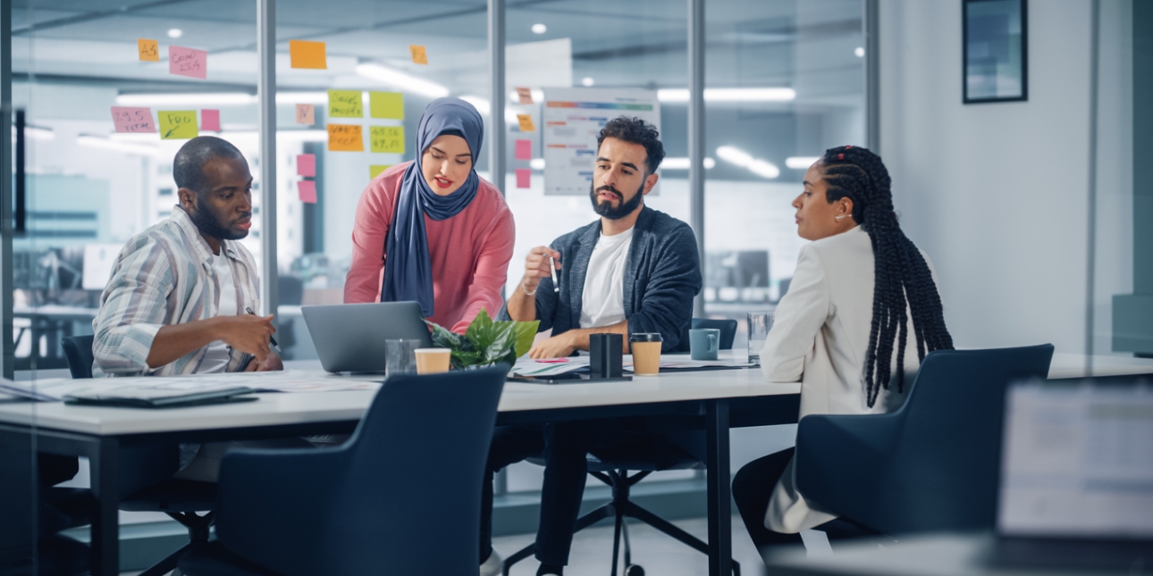 A woman in a blue hijab and pink jumper is stood pointing to a laptop while two male and one female colleague are sat around a board table