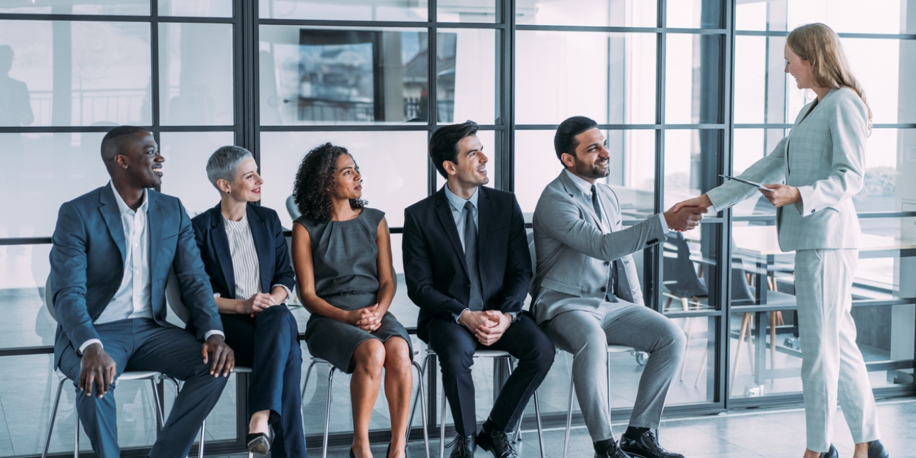 Five job candidates sit in a line against a glass office wall. There are two women and three men of different ethnicities. The man on the right shakes hand with the woman recruiter who is standing. All are smiling.