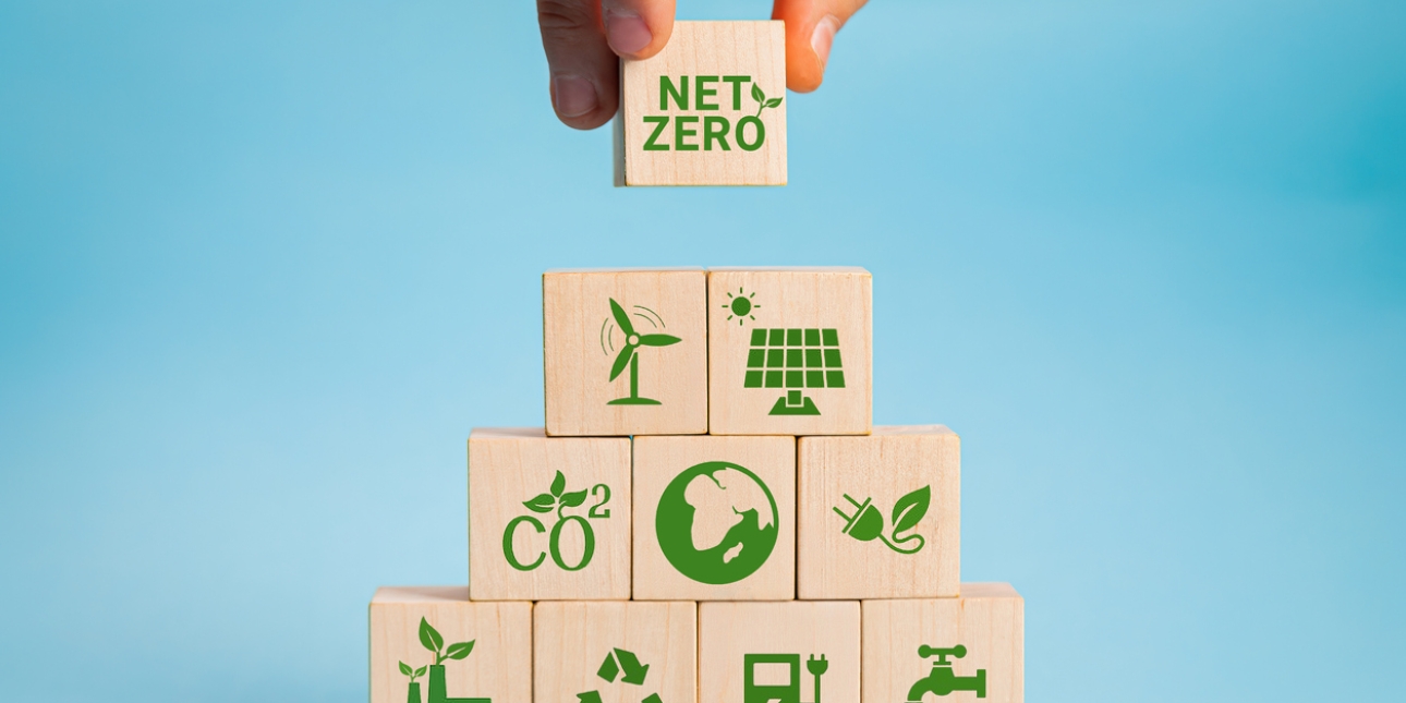 A hand places a wooden block featuring the words net zero, on top of a pyramid of similar blocks. These blocks each contain a word or icon representing: wind turbine, solar panel, CO2, a globe, green electricity, a green power station, recycling, ele