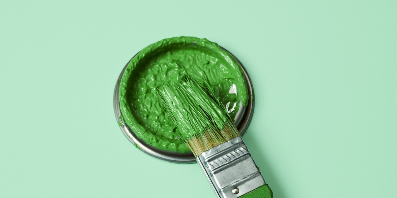 A paintbrush covered in green paint rests on the lid of a tin of green paint