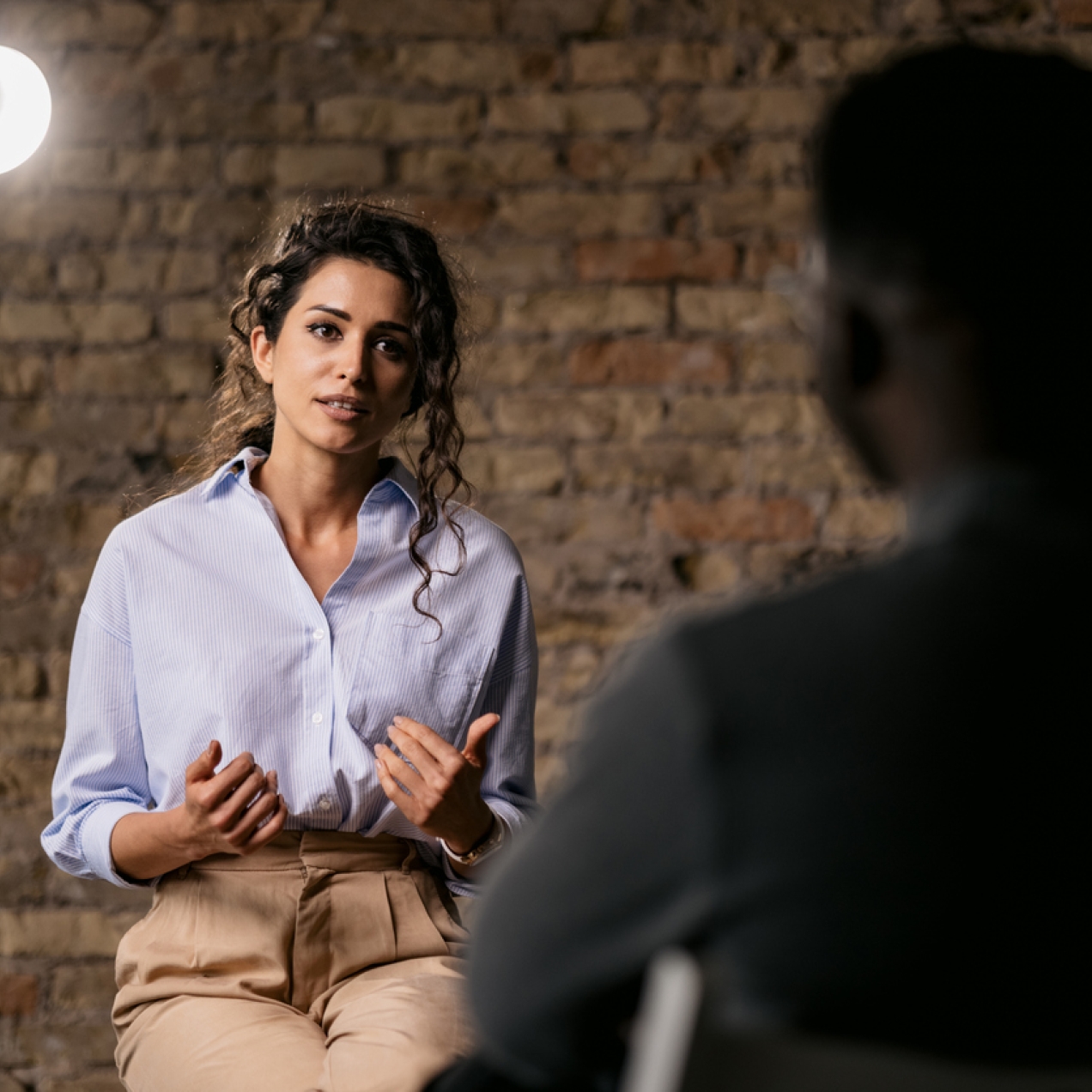 A woman under a spotlight is giving an interview to a journalist. Behind her is a brick wall.