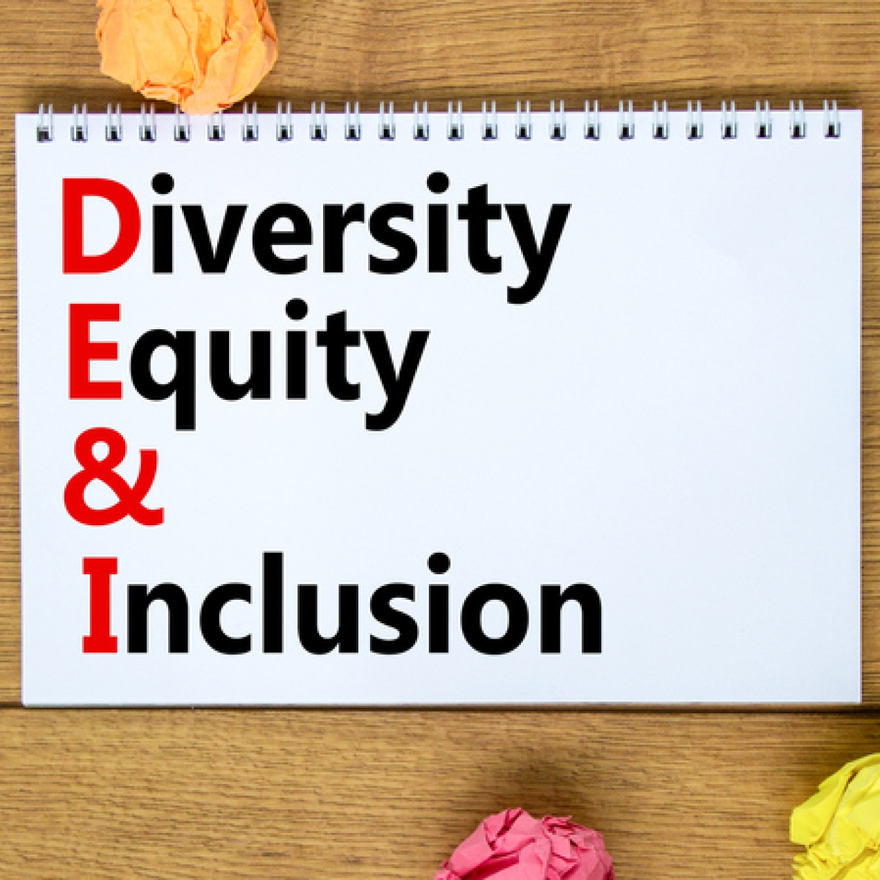 The words Diversity Equity & Inclusion printed on a notebook. To the left of the notebook is a black pen. Surrounding the notebook are four balls of paper in orange, turquoise, yellow and pink.