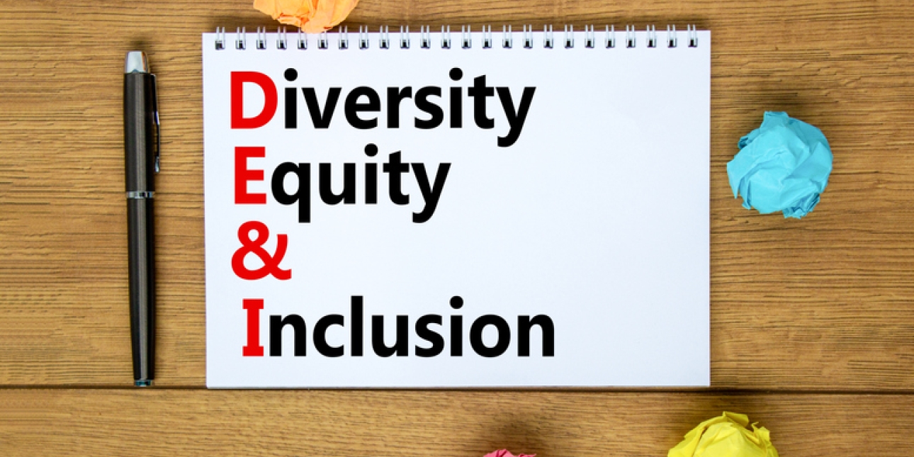 The words Diversity Equity & Inclusion printed on a notebook. To the left of the notebook is a black pen. Surrounding the notebook are four balls of paper in orange, turquoise, yellow and pink.