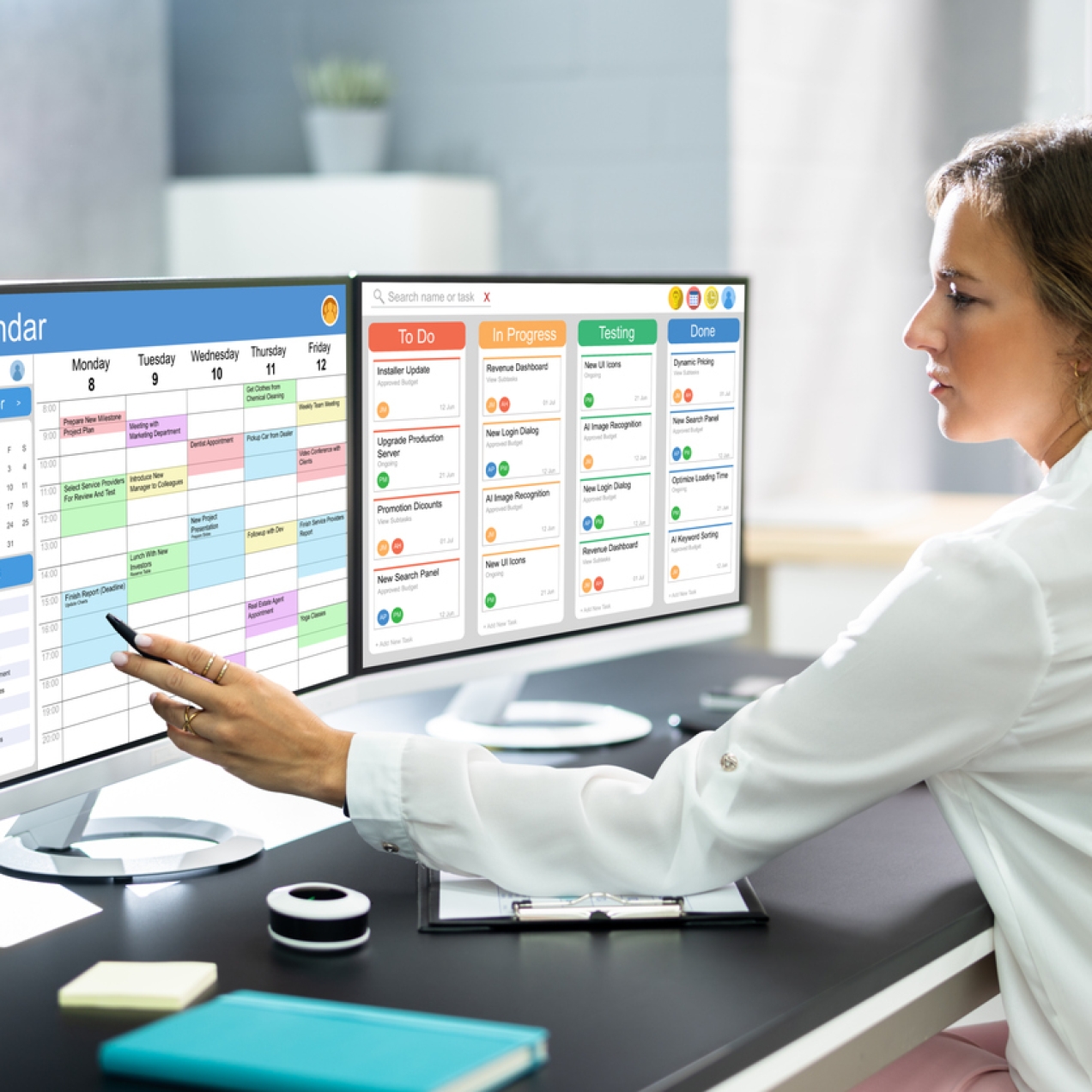 A white woman with blonde hair in a ponytail sits in front of two computer screens. She holds a pen at a colourful online calendar on the left. The right hand screen displays a schedule of tasks.