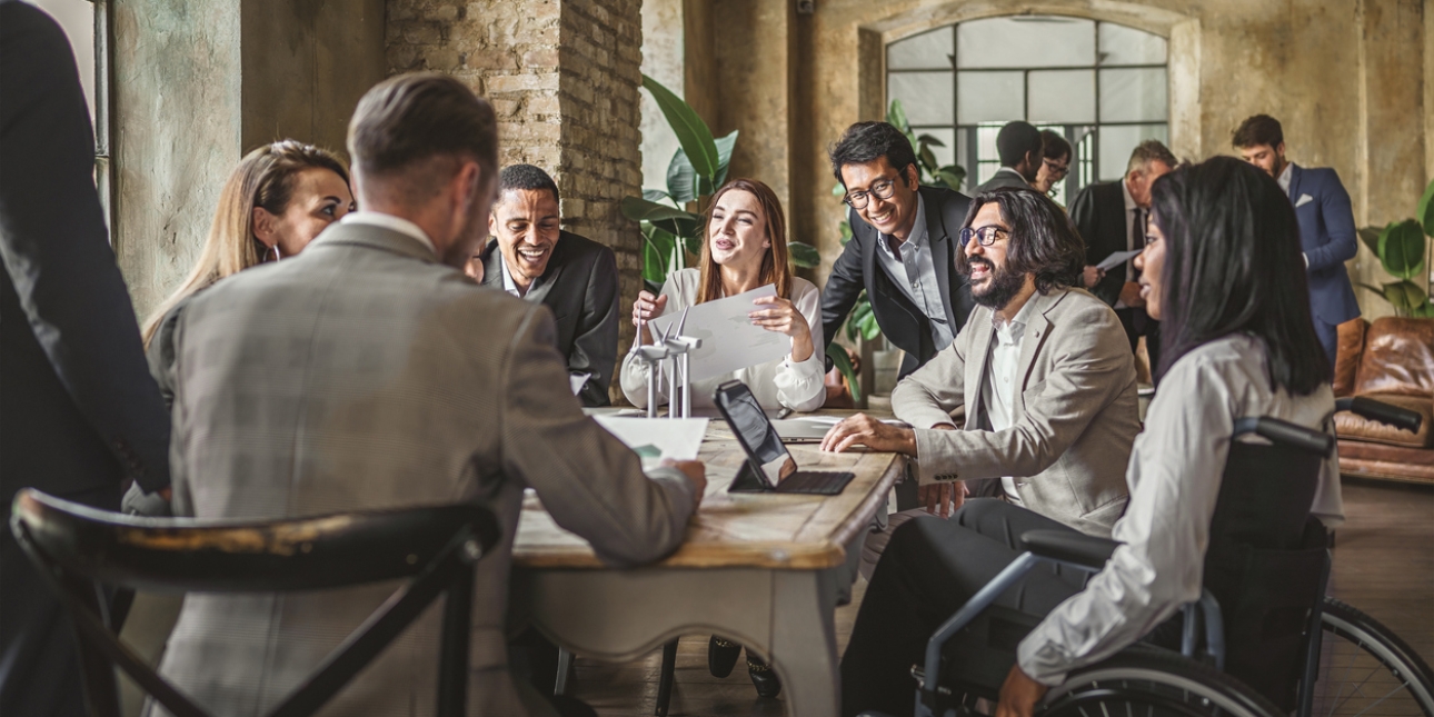 Six colleagues sat smiling around a table. One of the three women is in a wheelchair. Two colleagues are standing.