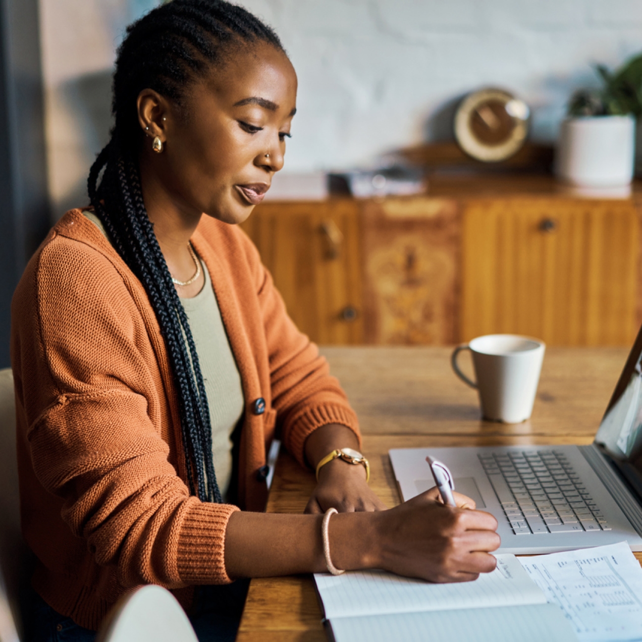 A Black woman with long hair and wearing an orange cardigan sits at a table writing notes in a book. In front of her is a laptop with four people on a webinar.