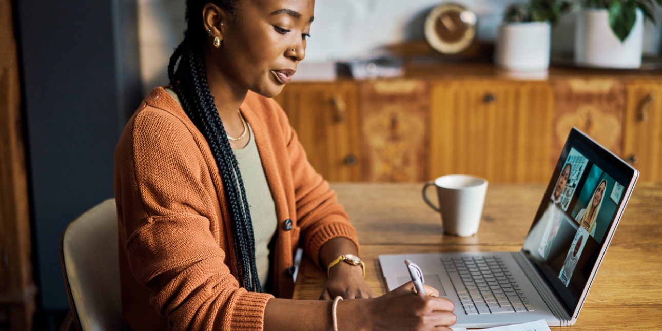 A Black woman with long hair and wearing an orange cardigan sits at a table writing notes in a book. In front of her is a laptop with four people on a webinar.