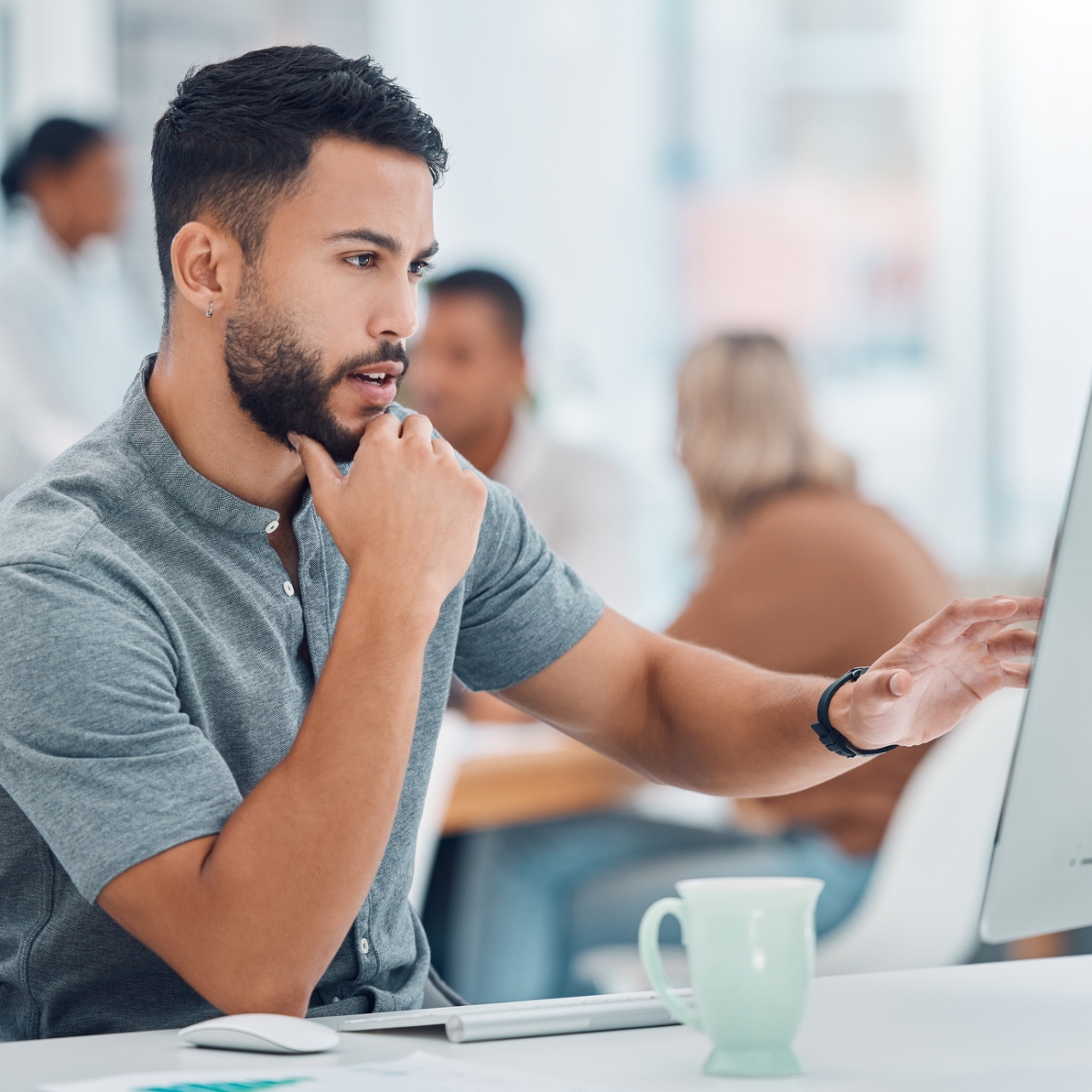 Dark haired man with beard wearing a grey short sleeved shirt looks at a computer monitor while sat in the office