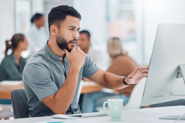 Dark haired man with beard wearing a grey short sleeved shirt looks at a computer monitor while sat in the office