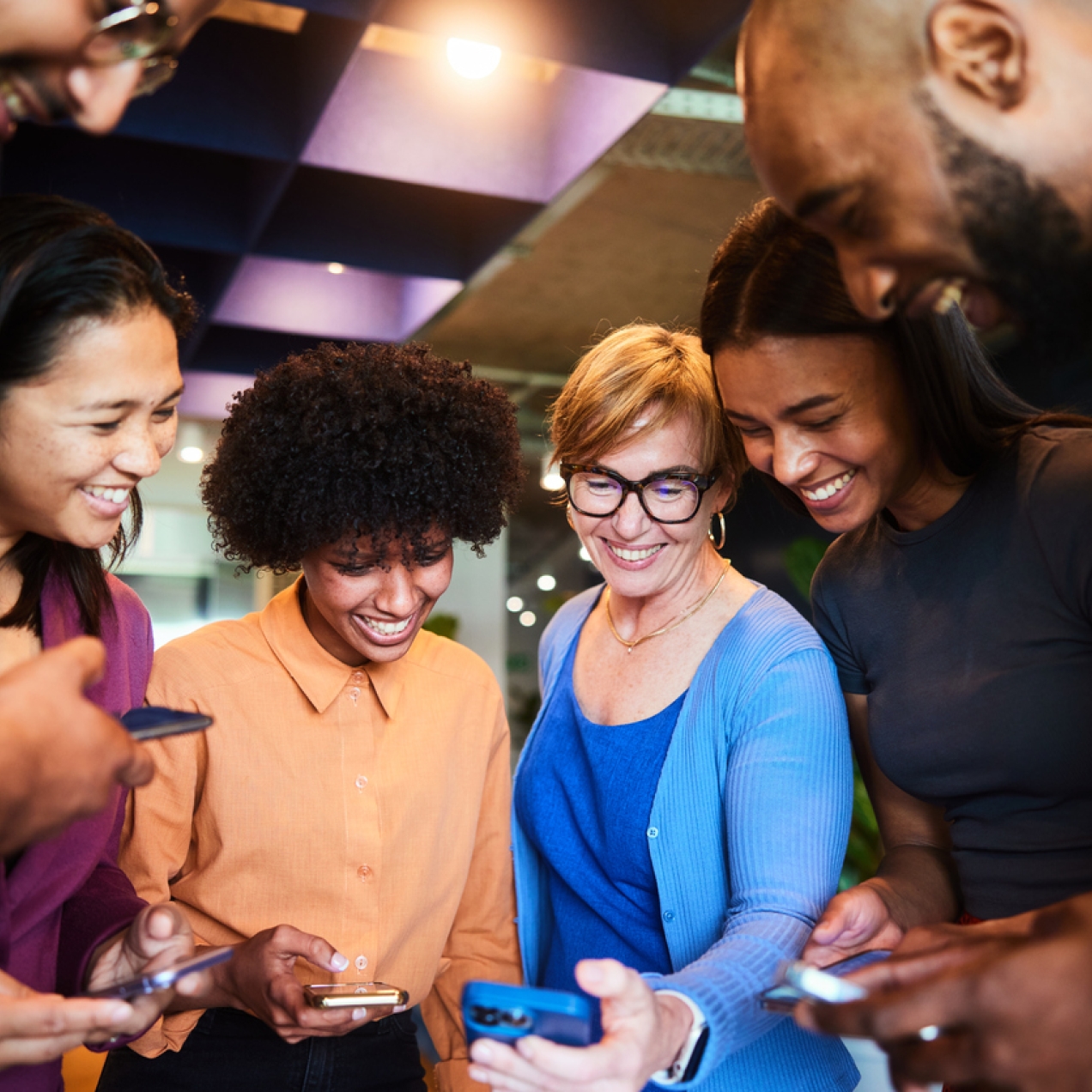 Four women and two men of different ethnicities look at each other's phones smiling at the screens