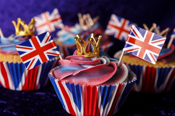 A selection of red, white and blue cupcakes with miniature gold crowns on top. There are assorted miniature two union (jack) flags stuck in each cake.