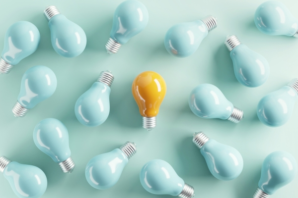 A bird's eye view of a yellow tinted light bulb laid flat on a teal background, surrounded by 15 turquoise-tinted bulbs