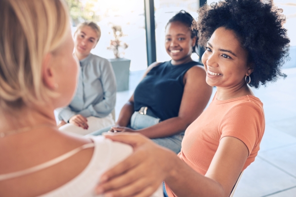 Black woman comforting a white colleague by putting a hand on her shoulder while sat round in a circle with other people