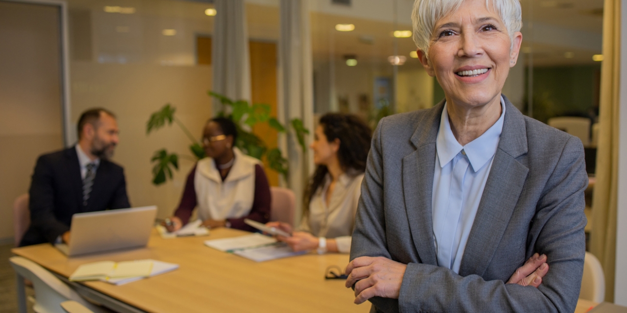 A white middle aged woman with grey hair, wearing a grey suit, stands with her arms cross in a boardroom. A white man, black woman and white woman are sat in the background.