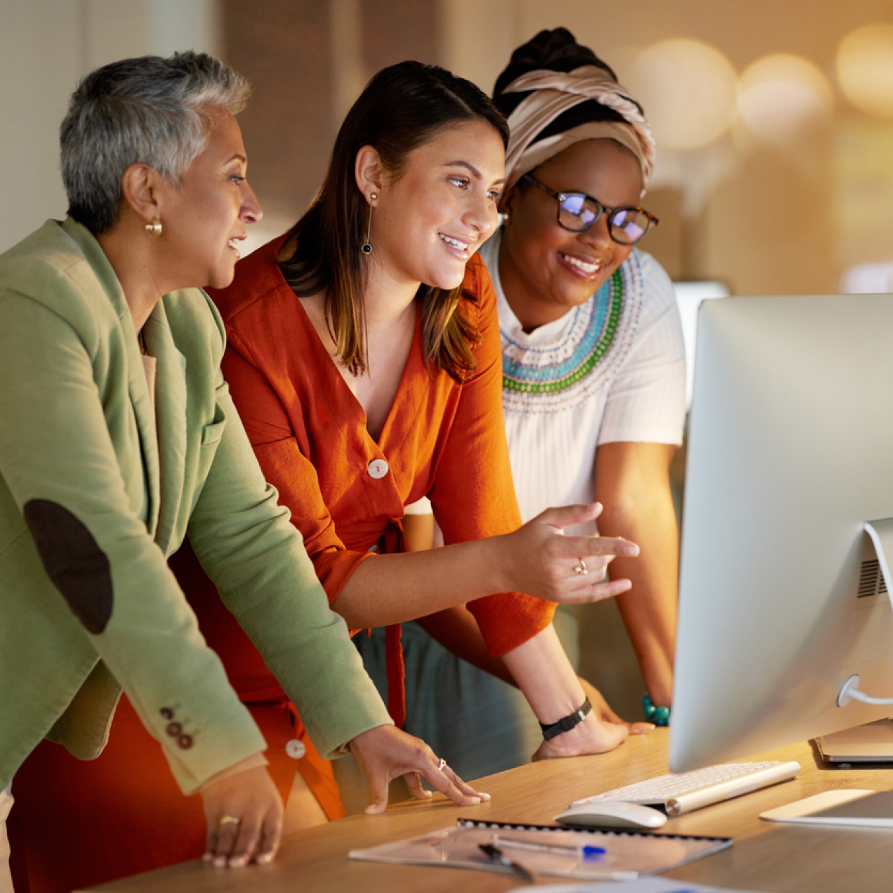 Three women of different ages and ethnicities lean over a desk looking at a computer monitor