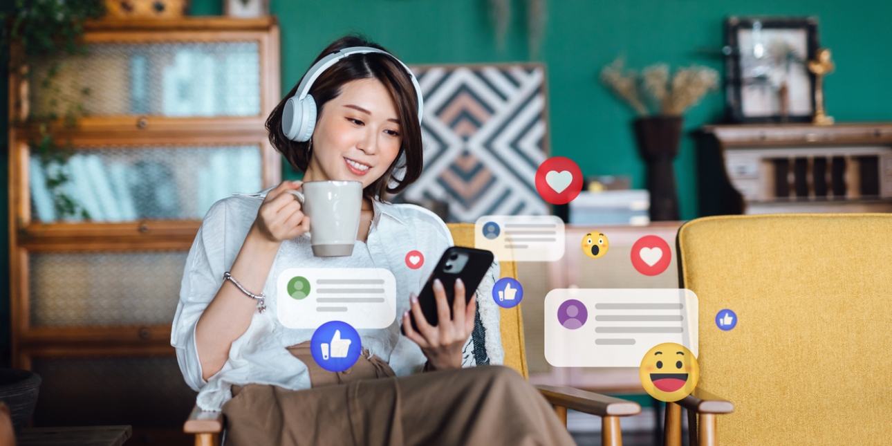 A young Asian woman is sat in a living room chair wearing headphones, with her phone in one hand and a cup in the other. Overlaid on the photo are digital illustrations of messages, emojis, hearts and thumbs up icons