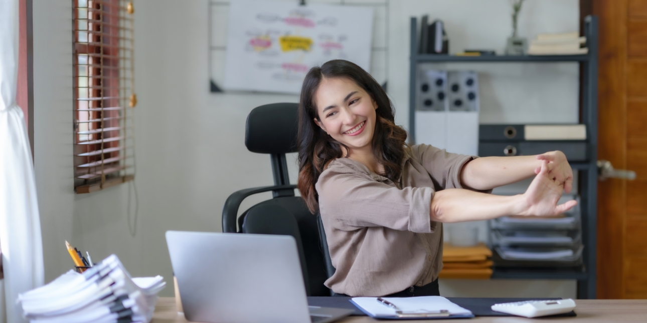 A smiling Asian business woman sat at her computer clasps both hands together while stretching her arms
