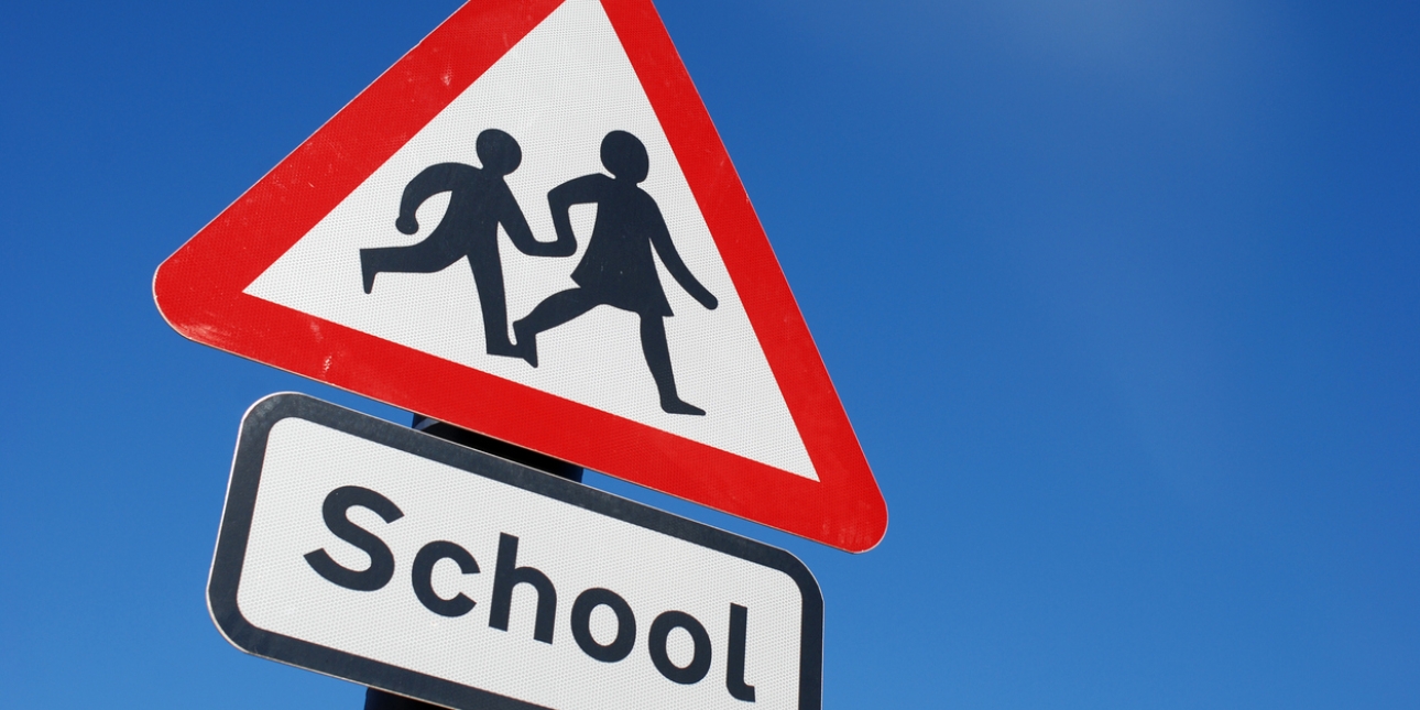 A British traffic sign showing school ahead. It is formed of a red triangle with a white background the silhouettes of two children. The word school appears on a plate underneath. The sky is a deep shade of blue.