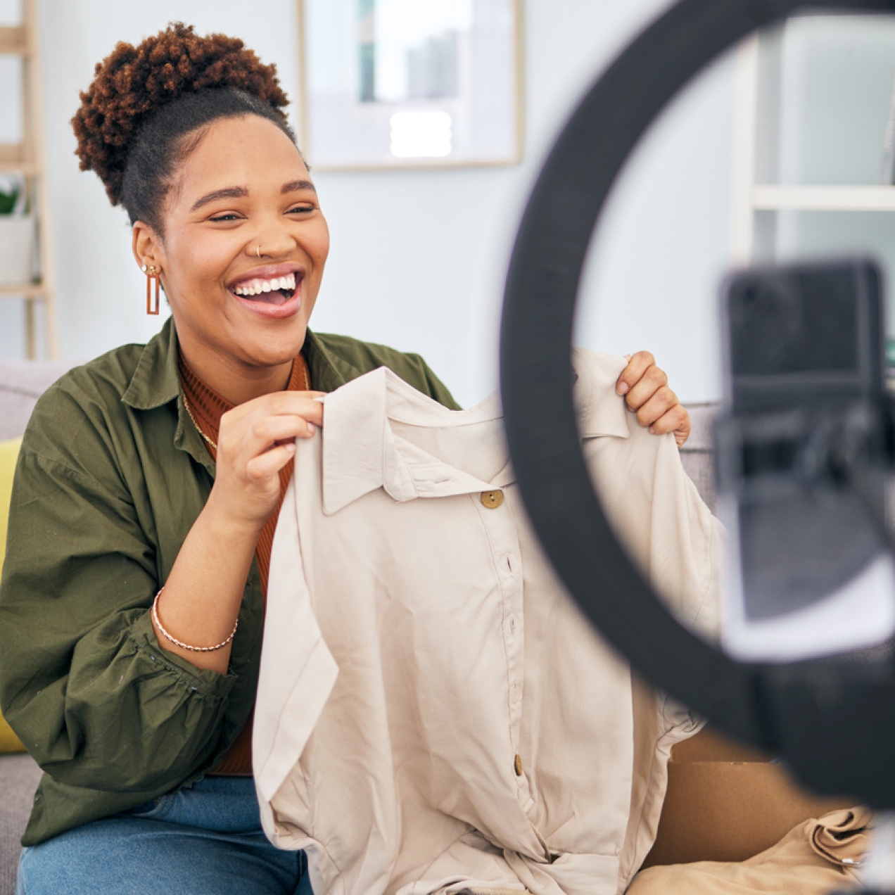 A black woman, who is sat on a sofa, laughs while holding a jacket in front of a mobile phone and ring light. In the background are a cushion and plants, books and a clock on a wooden shelving unit.