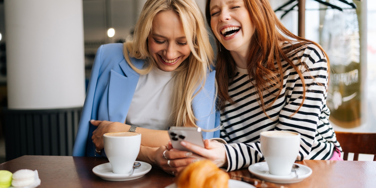 Two women, both white - one with blonde hair, the other with red hair, laughing while looking at a mobile phone screen. They are sat in a café.