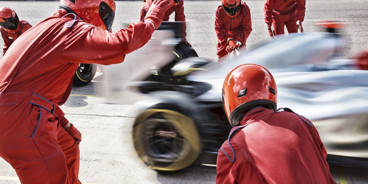 A team of six racing engineers, dressed in red overalls and red crash helmets with visors pulled down, wave off a racing car from the pit stop