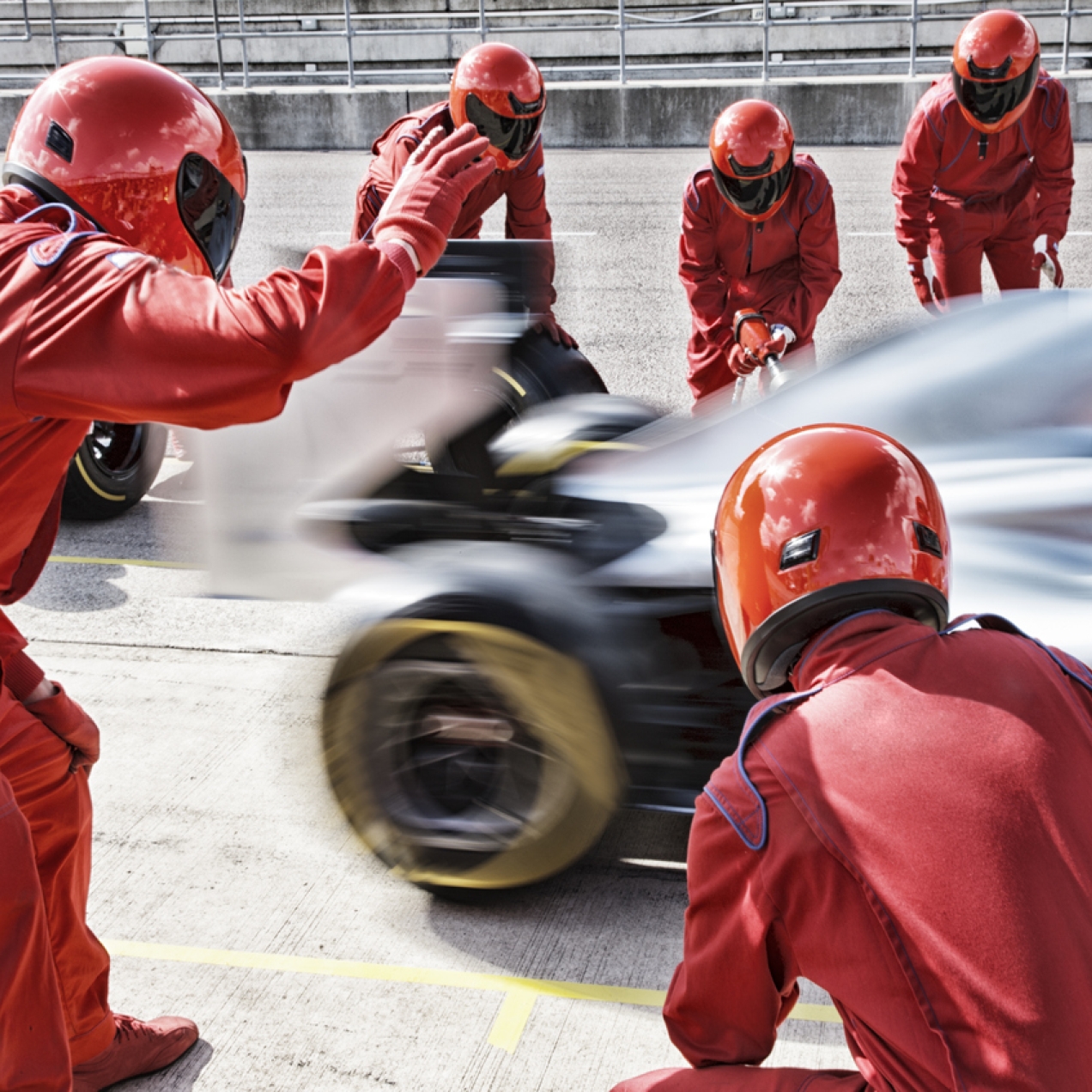 A team of six racing engineers, dressed in red overalls and red crash helmets with visors pulled down, wave off a racing car from the pit stop