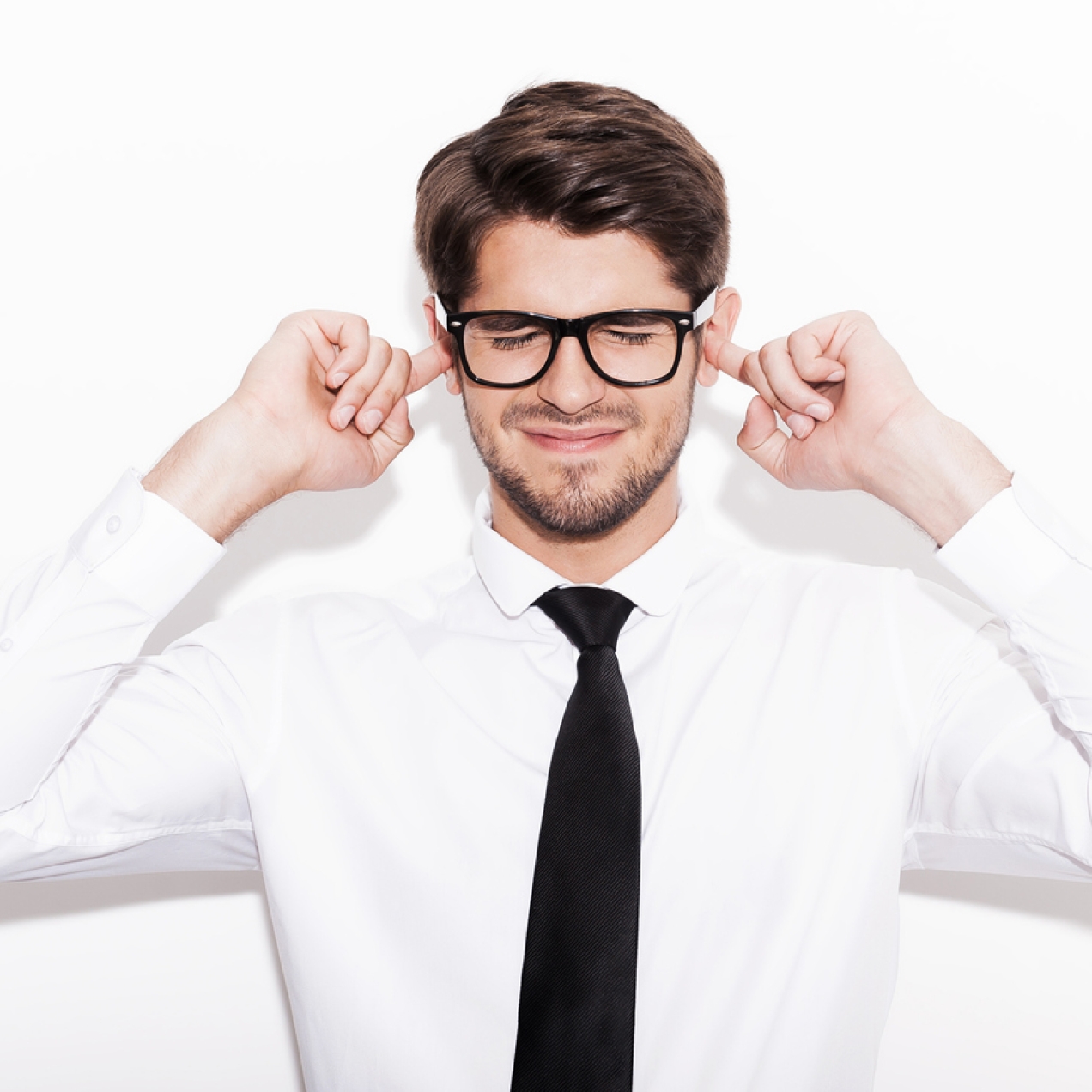 A white man with dark hair and thick rimmed glasses, wears a white long-sleeved shirt and black tie. His fingers are in his ears. The background is white