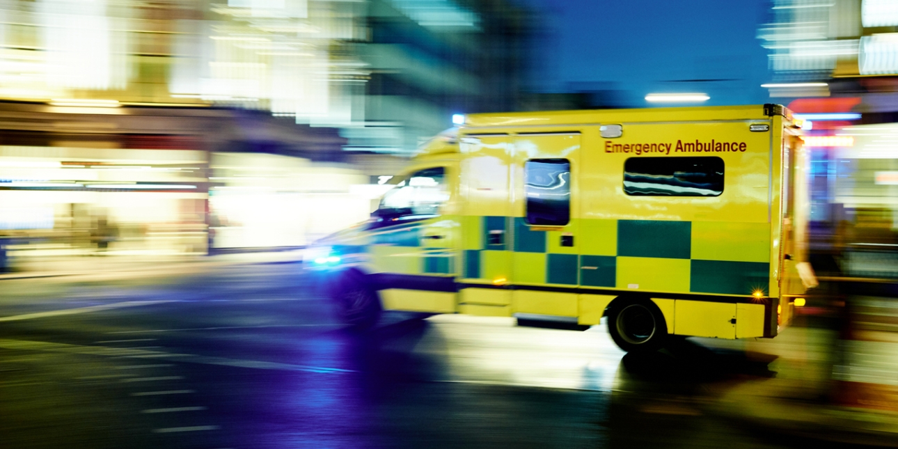 A yellow ambulance speeds through a British street. The background is blurred.