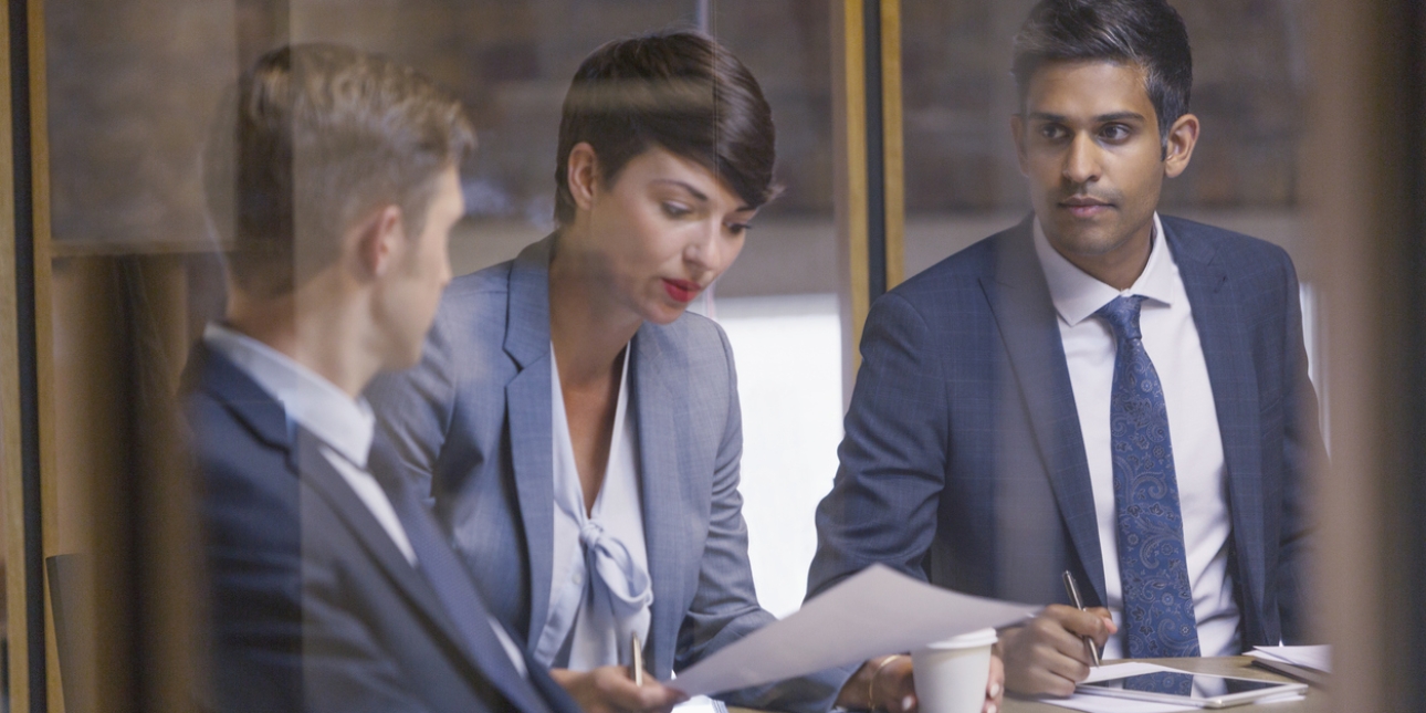 Three colleagues in business attire are sat in a meeting room. A white woman with dark hair is looking at paperwork. To her left is a white man with light brown hair wearing a grey suit. To the right is an Asian man with dark hair and blue suit and t