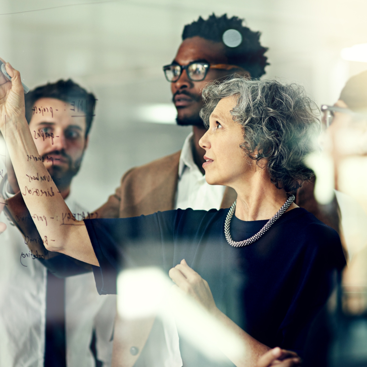 A business woman writes on glass with a marker while three male colleagues look on