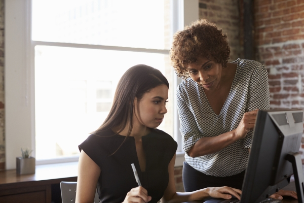A younger woman with a black top and dark hair is sat at a computer screen. Leaning over her and also looking at the screen is an older woman wearing a spotted blouse.