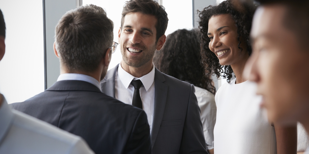 A white man with short dark hair, wearing a dark suit and tie, and a black woman with shoulder length curly hair and wearing a white blouse, smile at a business colleague at a networking event. The colleague has his back to the camera but appears in 