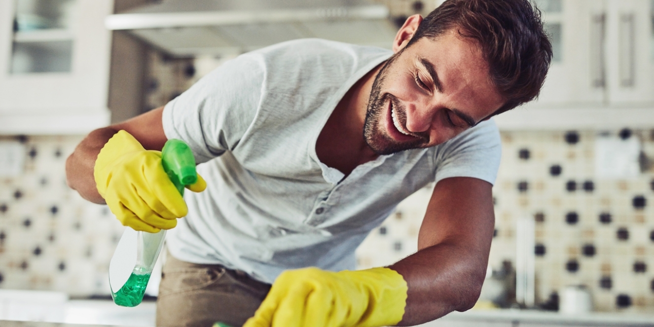 A white man with dark hair and wearing a t-shirt and yellow rubber globes holds a bottle of cleaning spray in his right hand and scrubs a kitchen surface with a scouring pad in his left hand. Behind him are kitchen units.