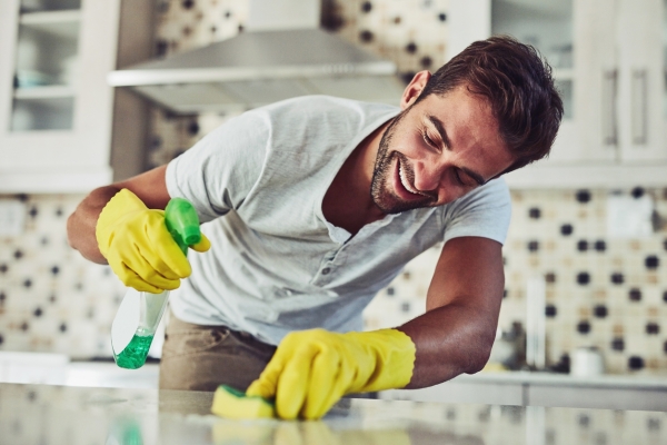 A white man with dark hair and wearing a t-shirt and yellow rubber globes holds a bottle of cleaning spray in his right hand and scrubs a kitchen surface with a scouring pad in his left hand. Behind him are kitchen units.