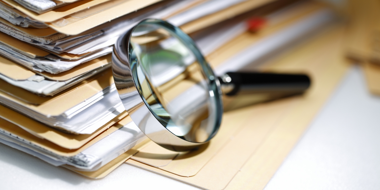 A magnifying glass rest on its side against a stack of folders containing paperwork