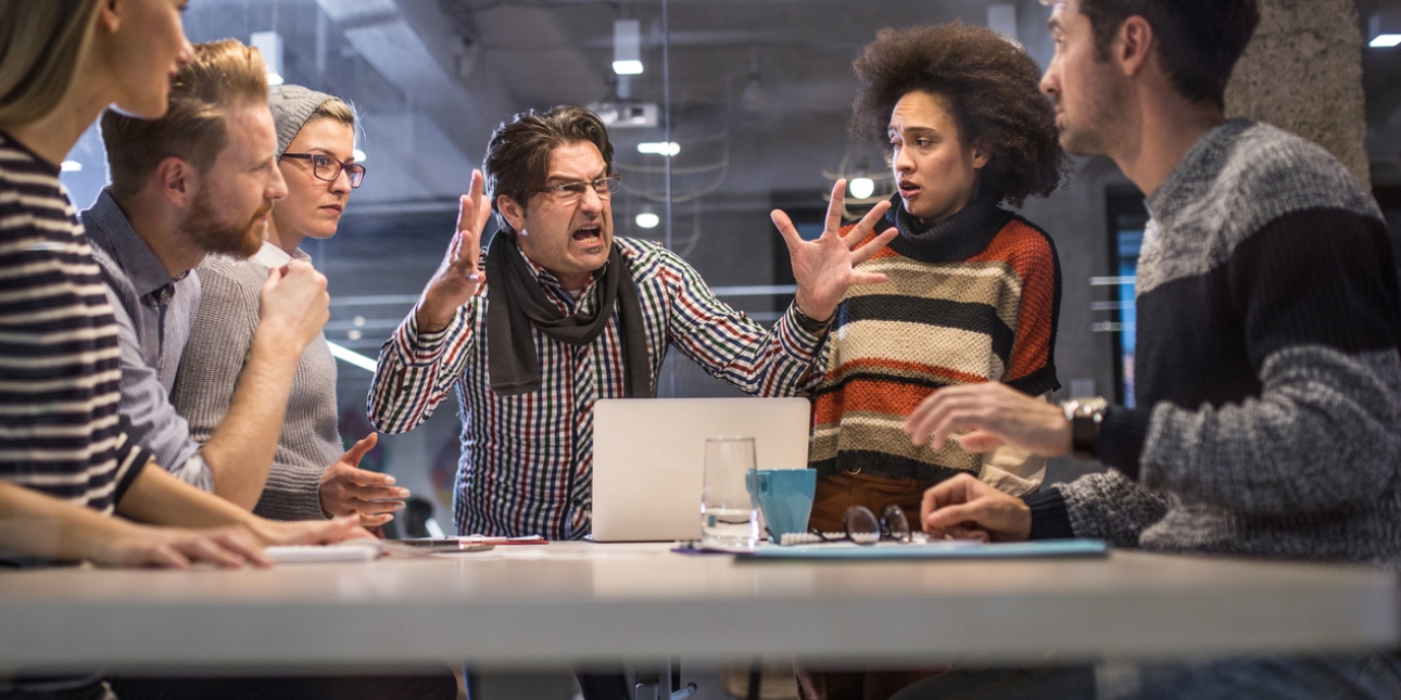 An angry looking boss raises both hands off the table, while his team of five looks on in shock with one wincing.