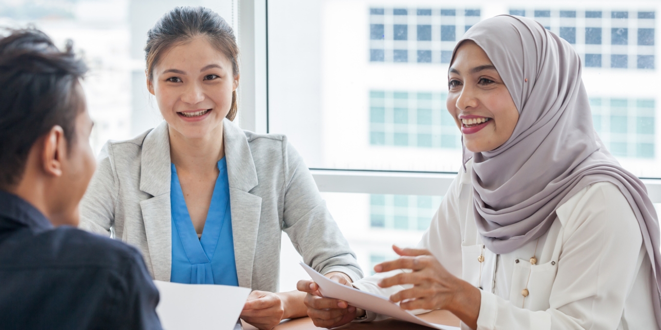 Two women interviewing a prospective employee. Both are smiling and wearing smart business clothes. The woman on the right wears a hijab. The interviewee is seen from the back.