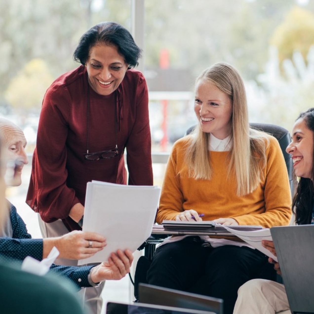 Eight smiling colleagues of different ages, genders and ethnicities gathered around a coffee table in discussion and looking at paperwork.